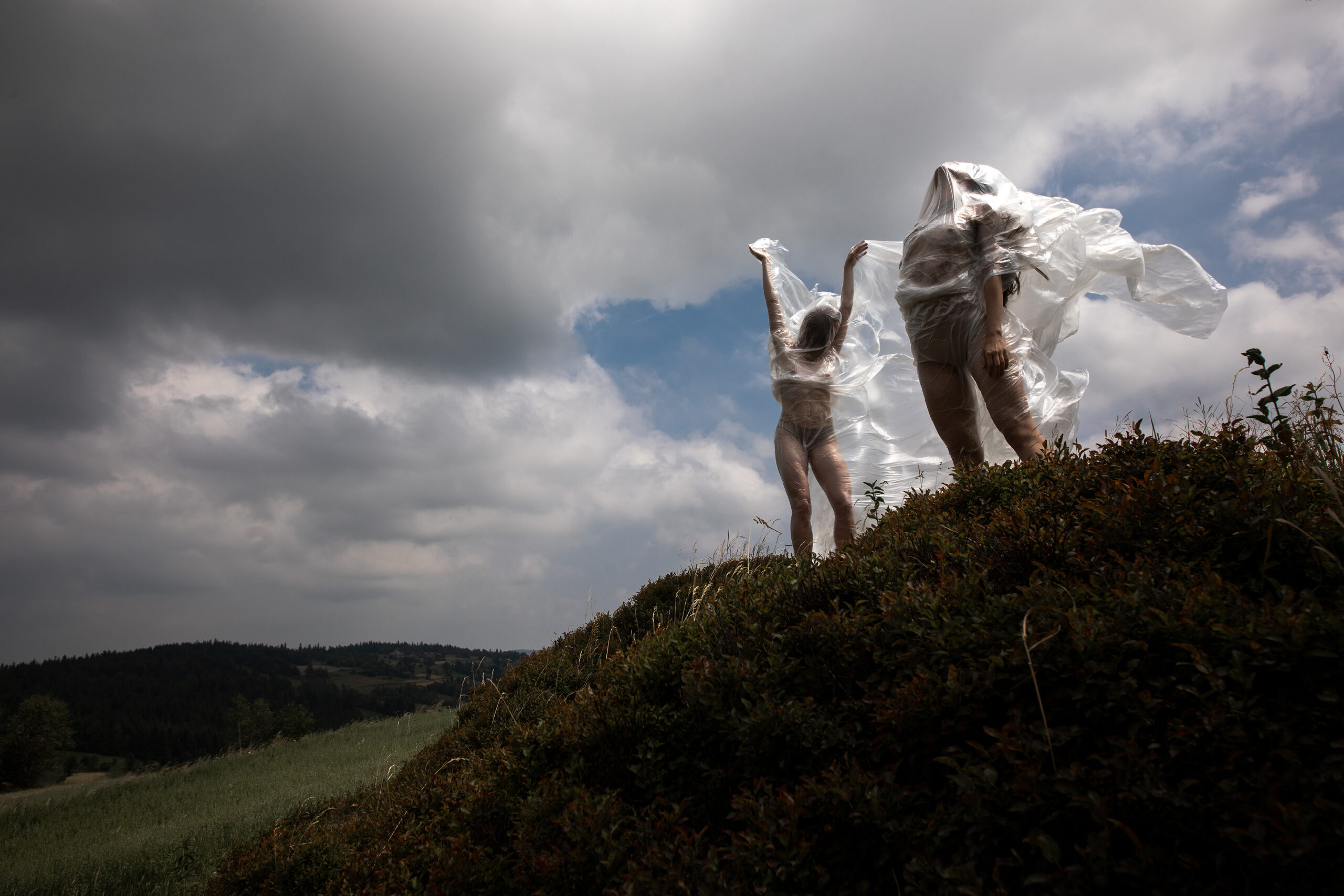 Dwie kobiety, stojące na wzgórzu, owinięte folią, która powiewa na wietrze. W tle chmury na niebie.