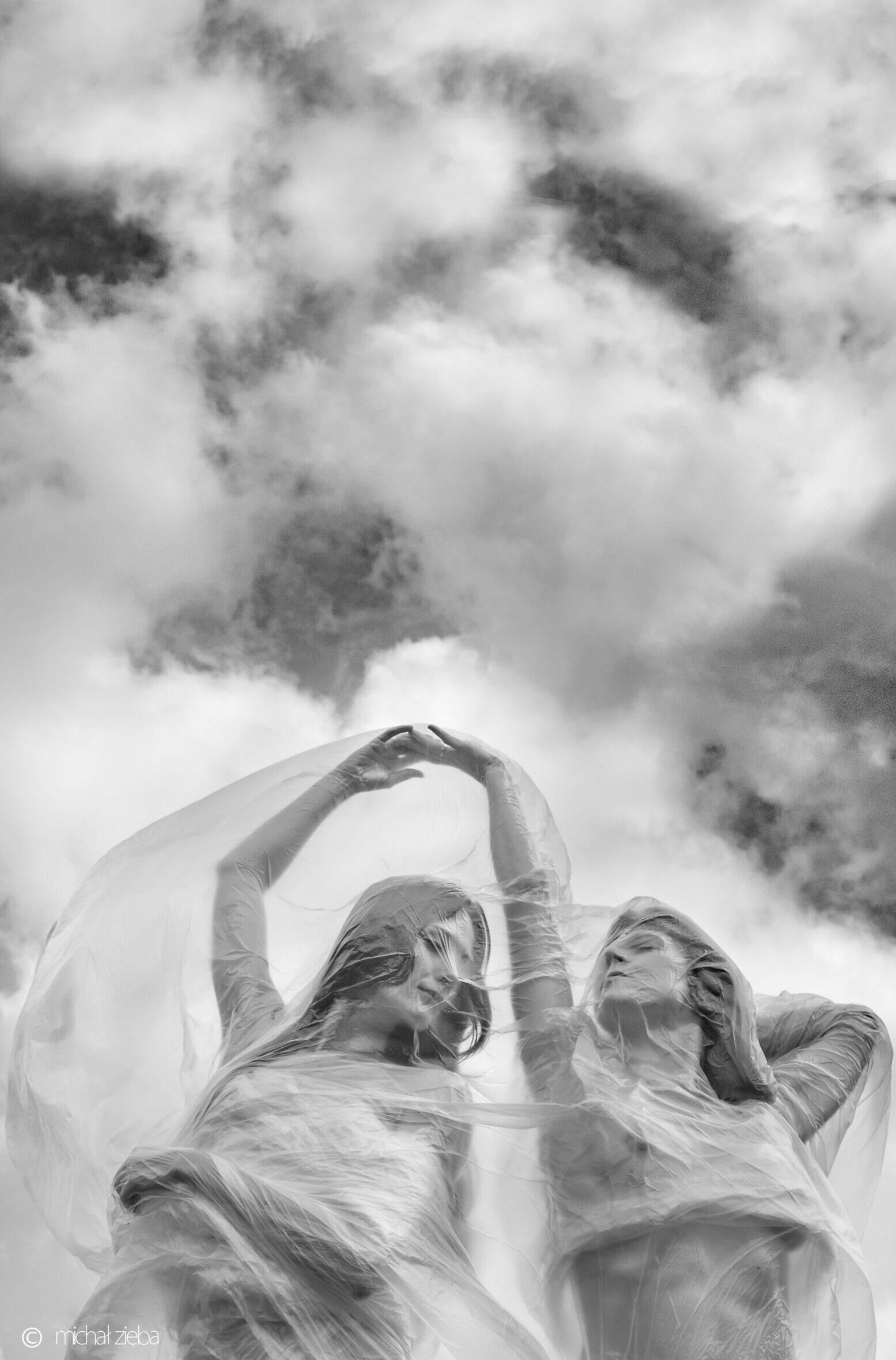Dwie kobiety owinięte folią, w tle majestatyczne, białe chmury. Zdjęcie w odcieniach szarości.
