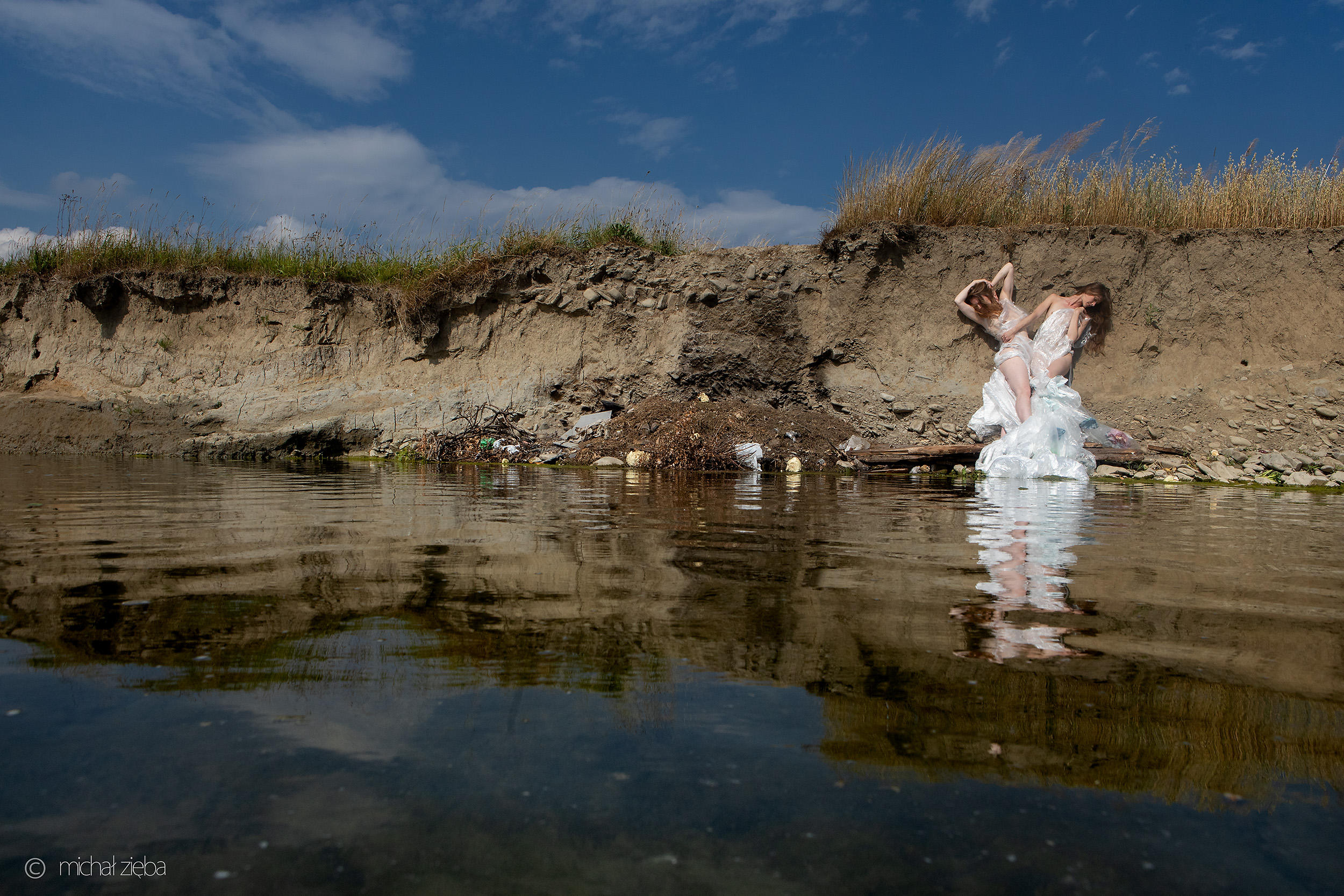 Dwie kobiety stojące pod skarpą brzegu rzeki, owinięte w jedną białą folię spływającą do wód rzeki.